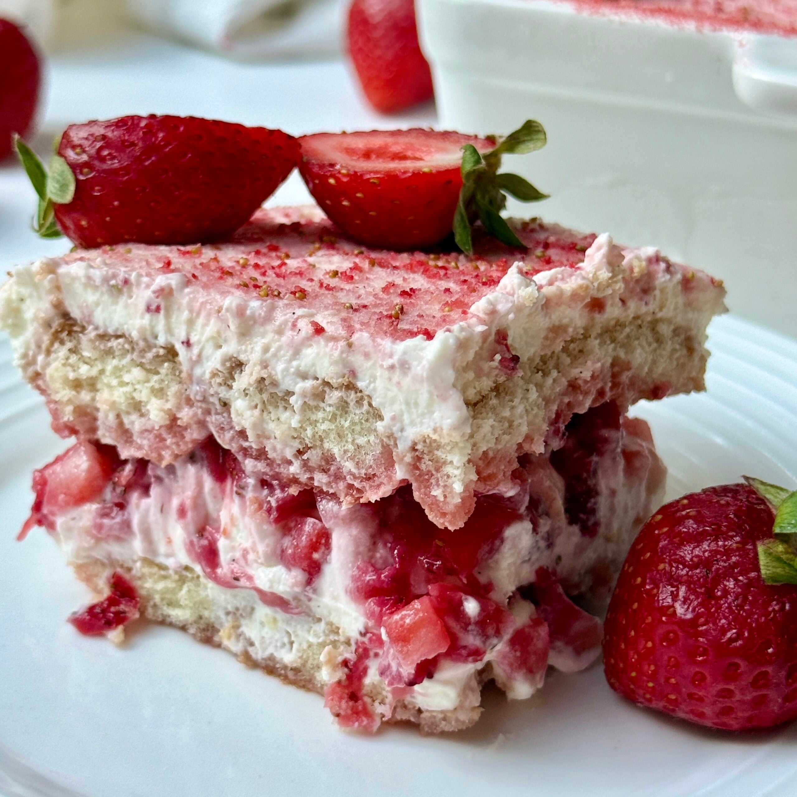 A slice of strawberry tiramisu layered with cream and strawberries sits on a white plate, garnished with a halved strawberry on top and a whole strawberry next to it. More strawberries and the dessert dish are in the background.