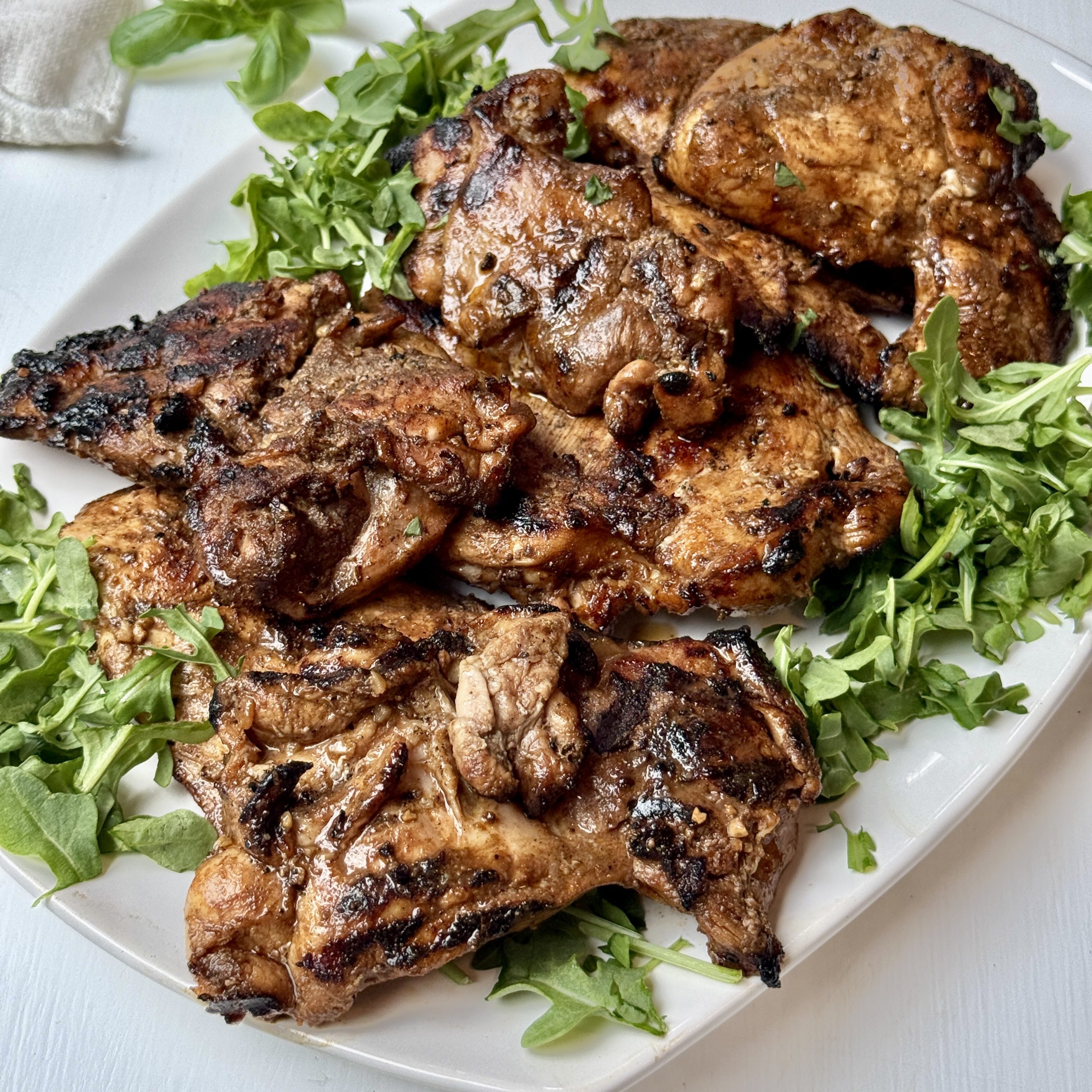 Grilled, marinated chicken thighs served on a white plate, garnished with fresh arugula leaves, on a light-colored surface.