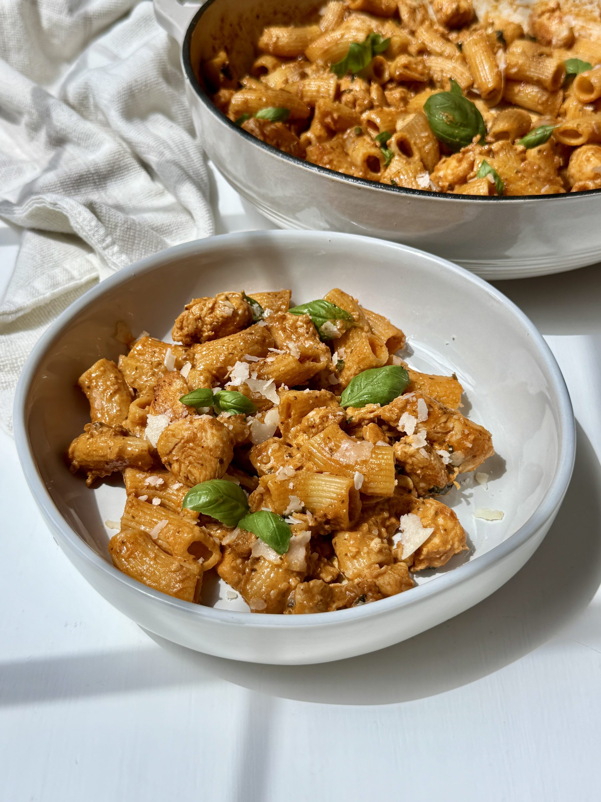 A bowl of red pesto pasta with chicken and the skillet in the background. 