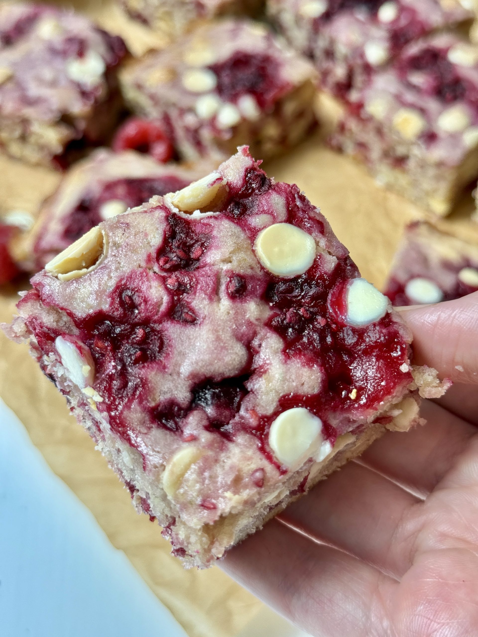 A hand holding up a white chocolate raspberry blondie with more on a table below.