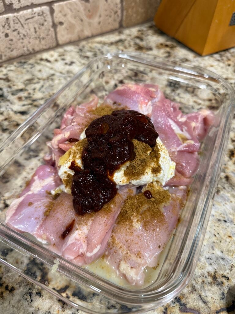 Raw chicken thighs in a glass dish are topped with a dollop of white yogurt, brownish spice, and a dark red sauce. The dish sits on a speckled granite countertop near a tan brick backsplash.