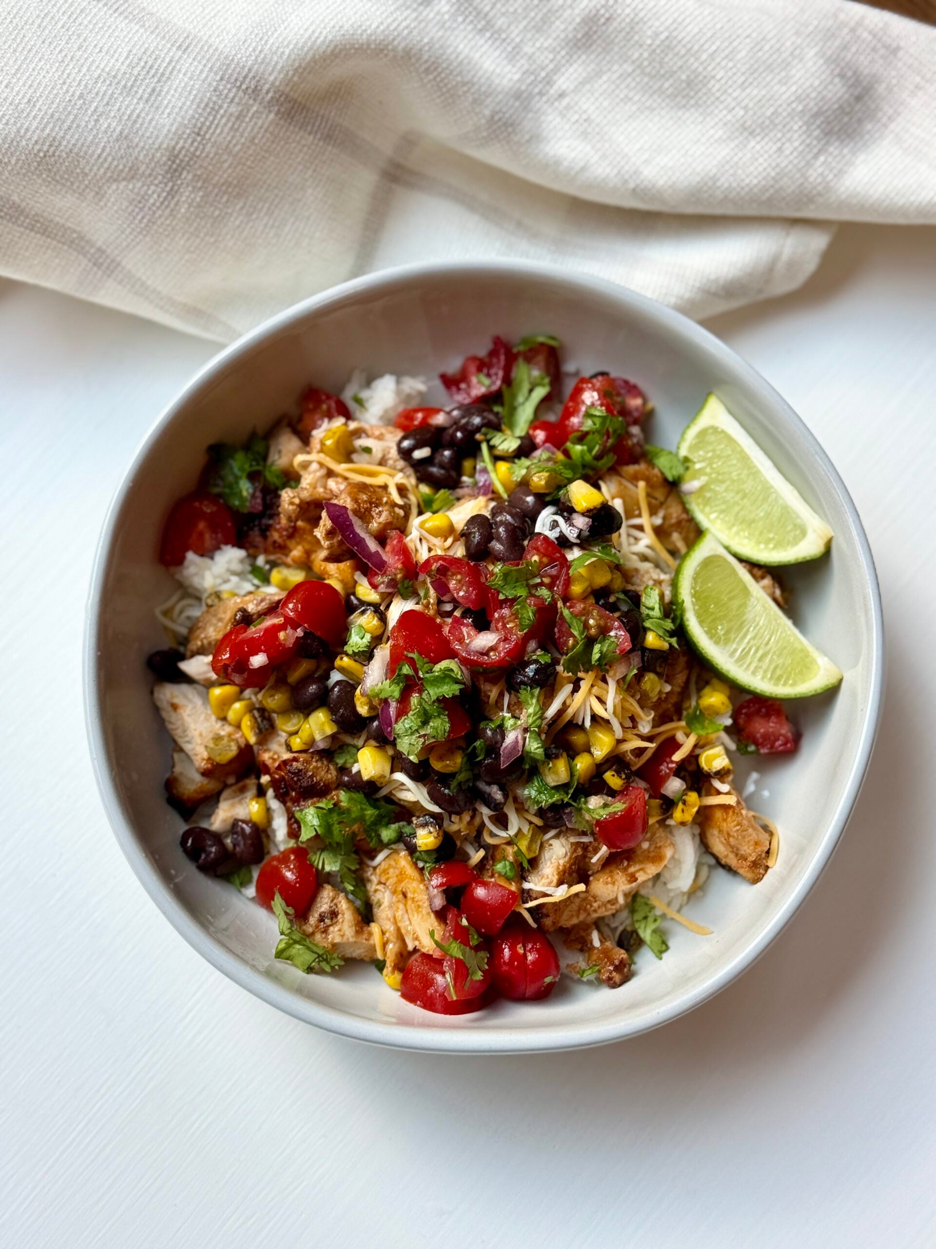 A bowl filled with rice, grilled chicken, black beans, corn, tomatoes, shredded cheese, cilantro, and two lime wedges.