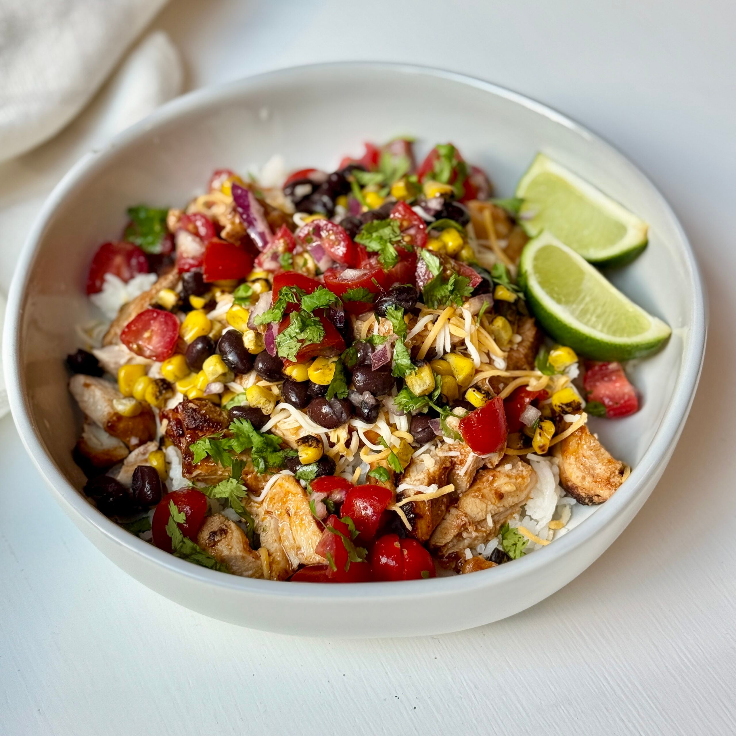 A bowl filled with rice, grilled chicken, black beans, corn, tomatoes, shredded cheese, cilantro, and two lime wedges.