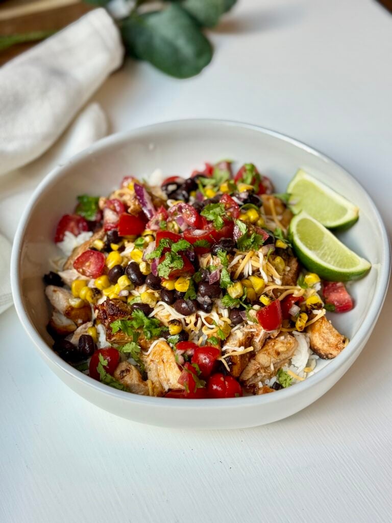 A bowl of rice topped with grilled chicken, black beans, corn, tomatoes, shredded cheese, and cilantro, garnished with two lime wedges on the side.
