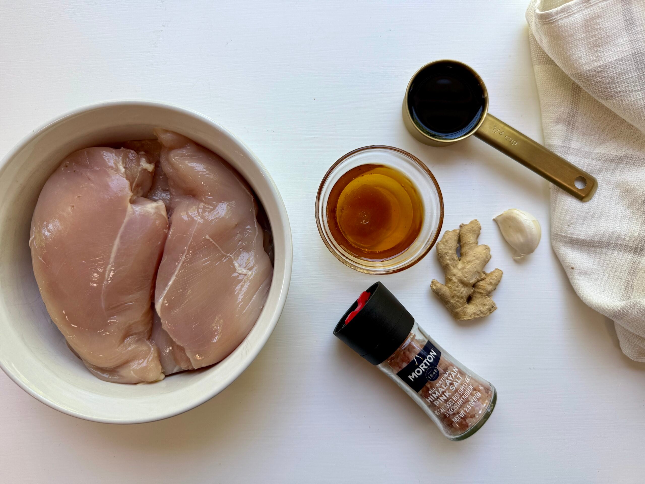Two raw chicken breasts in a bowl, with a cup of soy sauce, a small bowl of honey, a piece of ginger, a garlic clove, and a pepper grinder arranged on a white surface next to a white cloth.