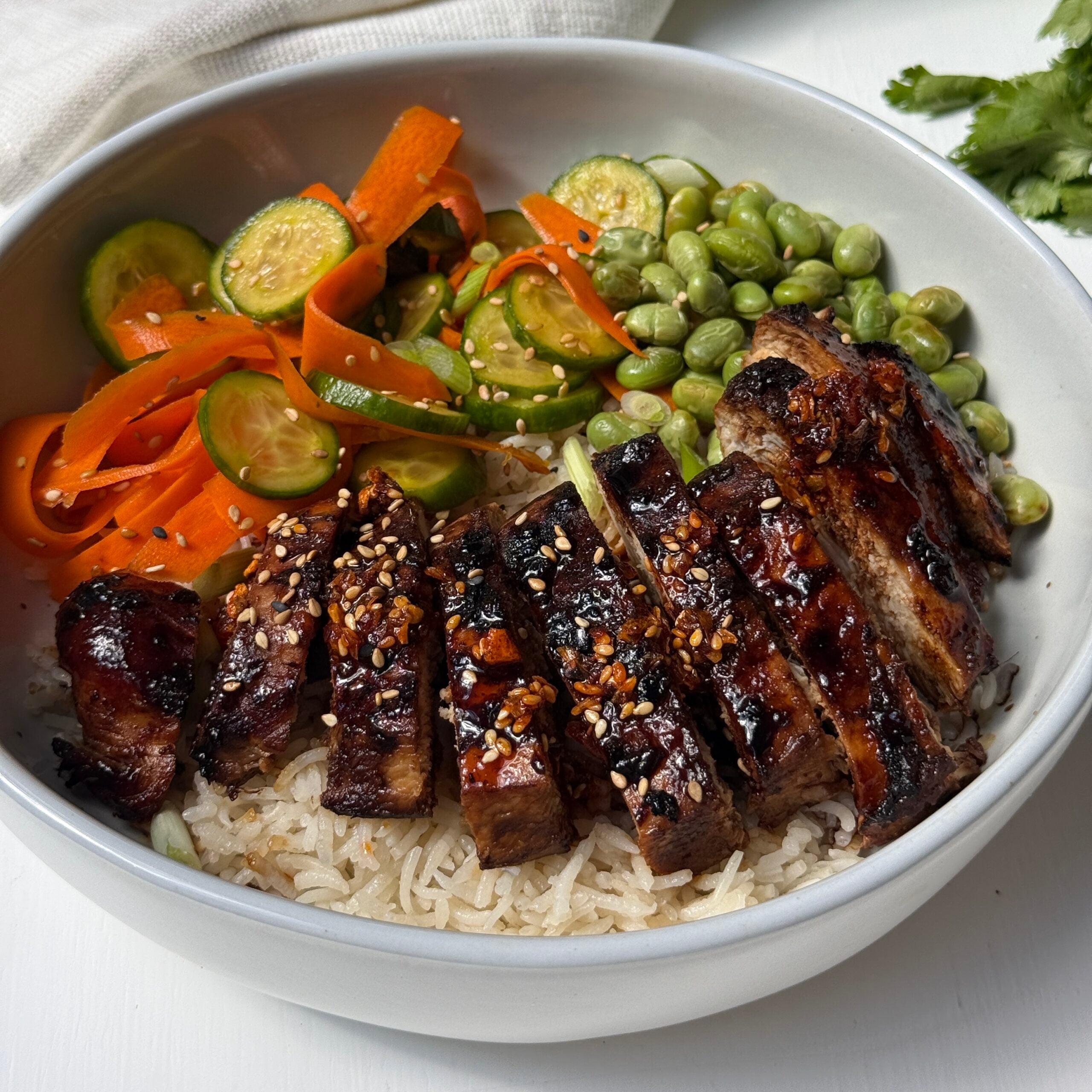 A bowl with grilled chicken on rice, served with edamame, sliced carrots, cucumber, and green onions. Garnished with sesame seeds. Scallions and cilantro are in the background.