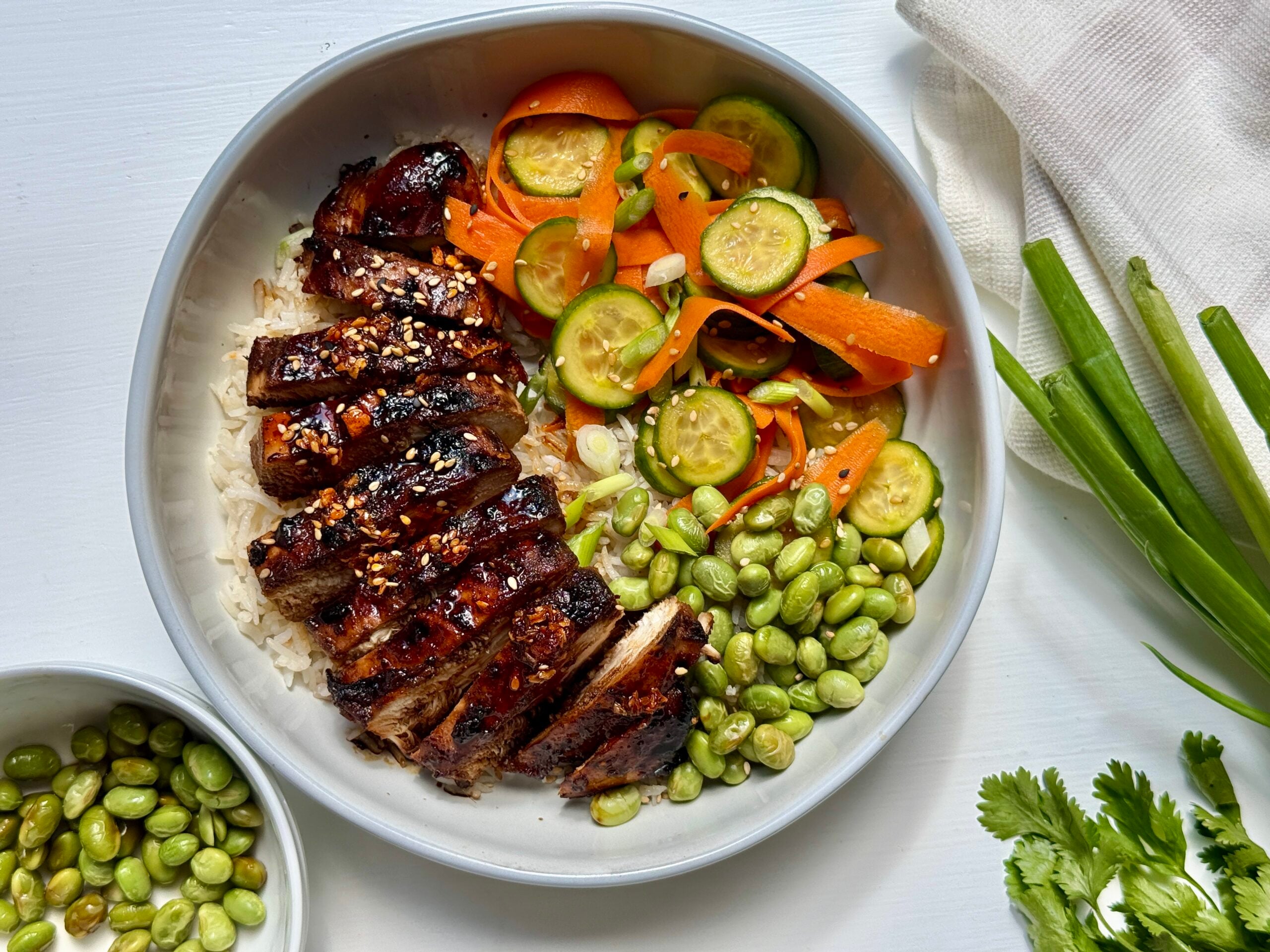 A bowl with sliced grilled chicken on rice, edamame, carrot and cucumber ribbons, and sesame seeds. Green onions and cilantro are nearby.