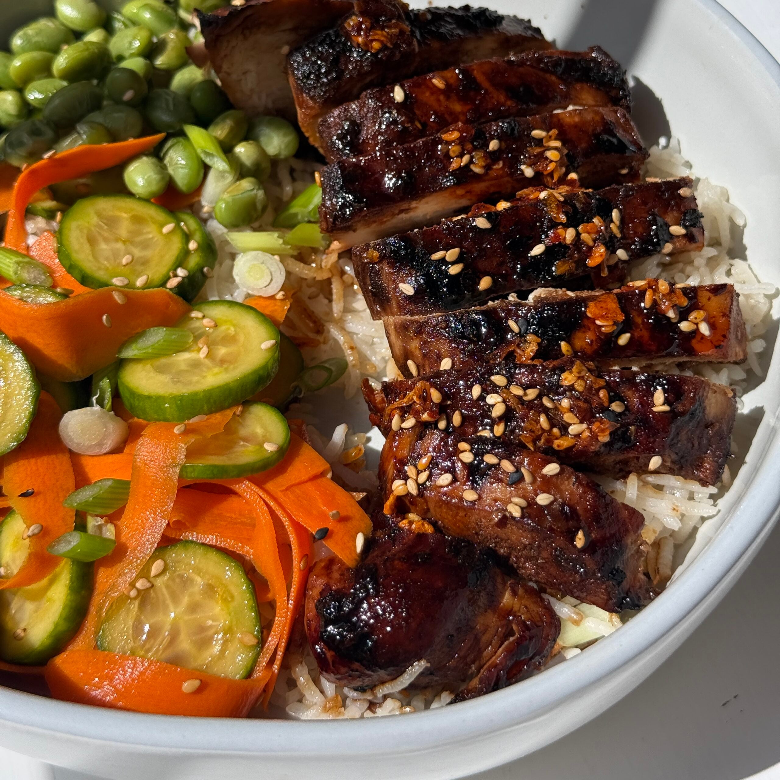 A bowl with grilled chicken on rice, served with edamame, sliced carrots, cucumber, and green onions. Garnished with sesame seeds.