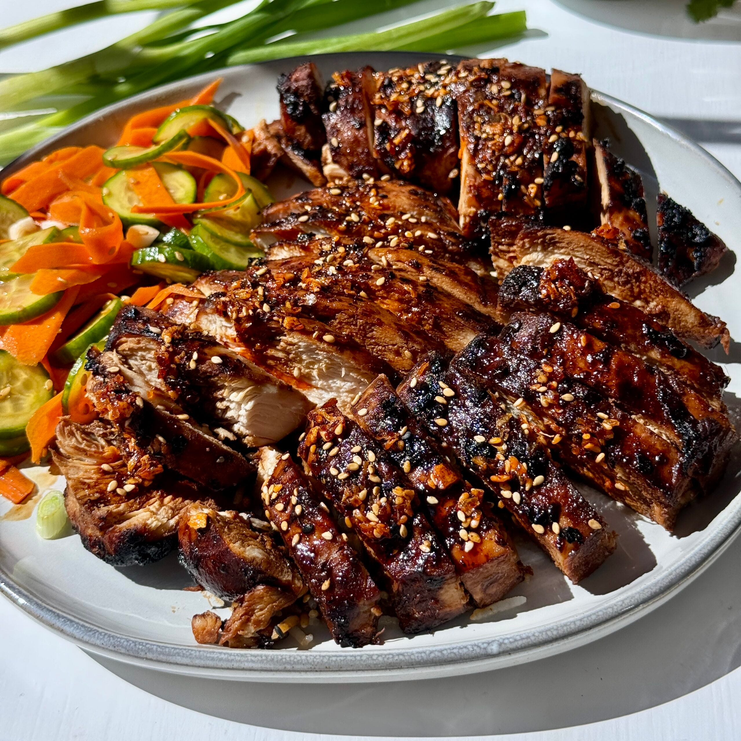 Sliced grilled meat with a shiny glaze, sprinkled with sesame seeds and spices, served on a white plate with a colorful side of sliced carrots, cucumbers, and green onions.