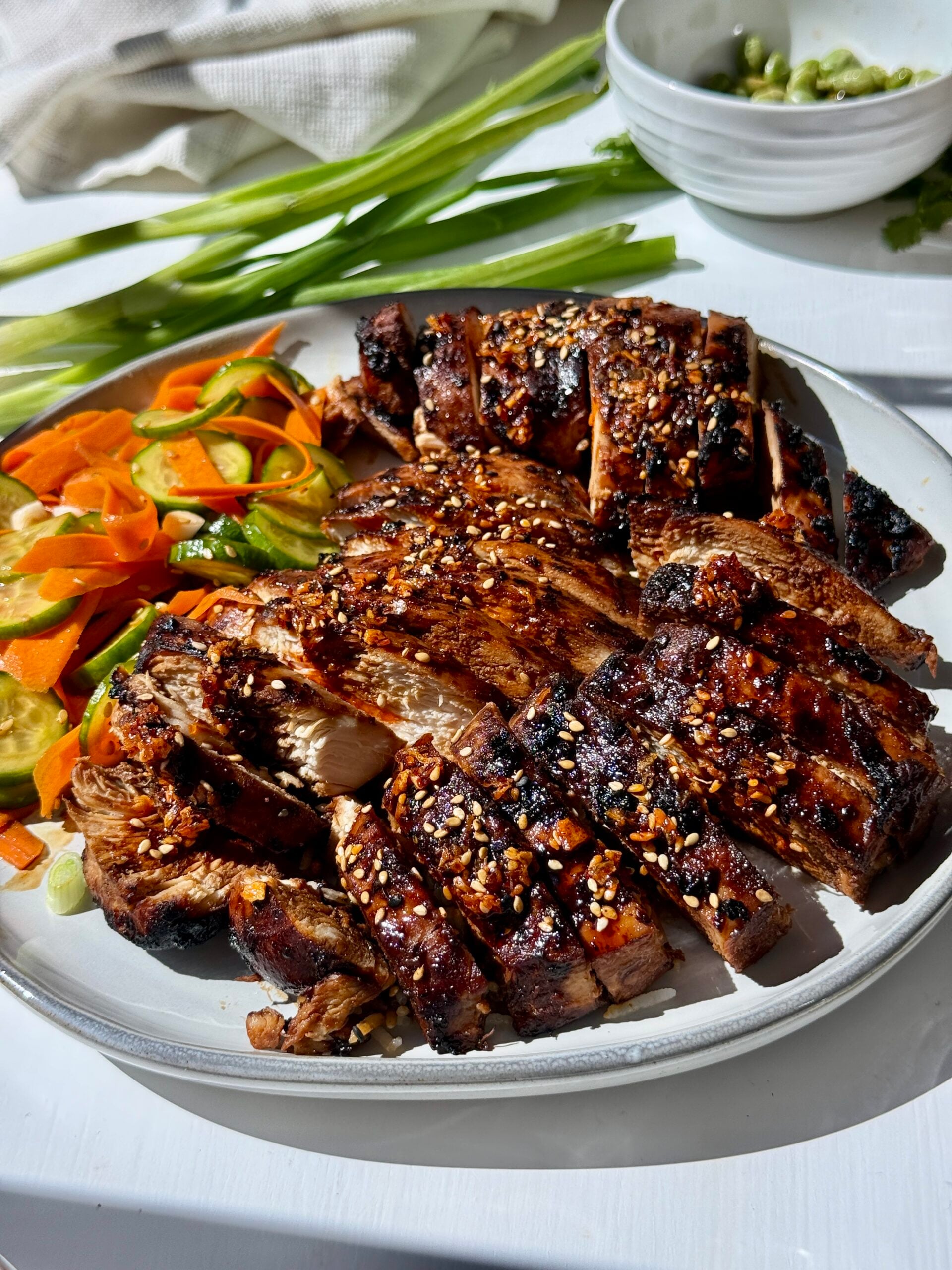 Sliced grilled chicken topped with sesame seeds and spices, served with a side of pickled vegetables and green onions on a white plate. Bright natural light enhances the dish.