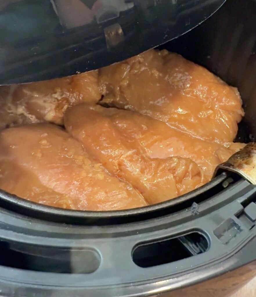 Four raw, marinated chicken breasts are placed inside an air fryer basket, ready to be cooked.