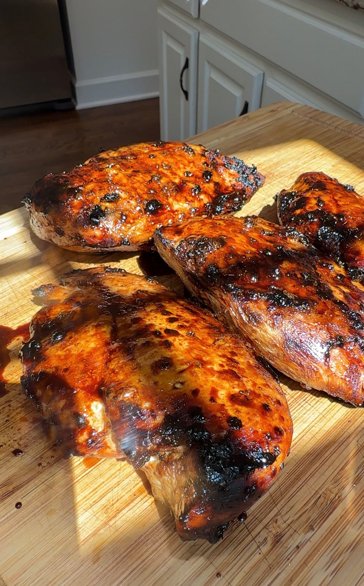 Four pieces of grilled, glazed chicken with char marks rest on a wooden cutting board in sunlight, showcasing caramelized, golden-brown skin.