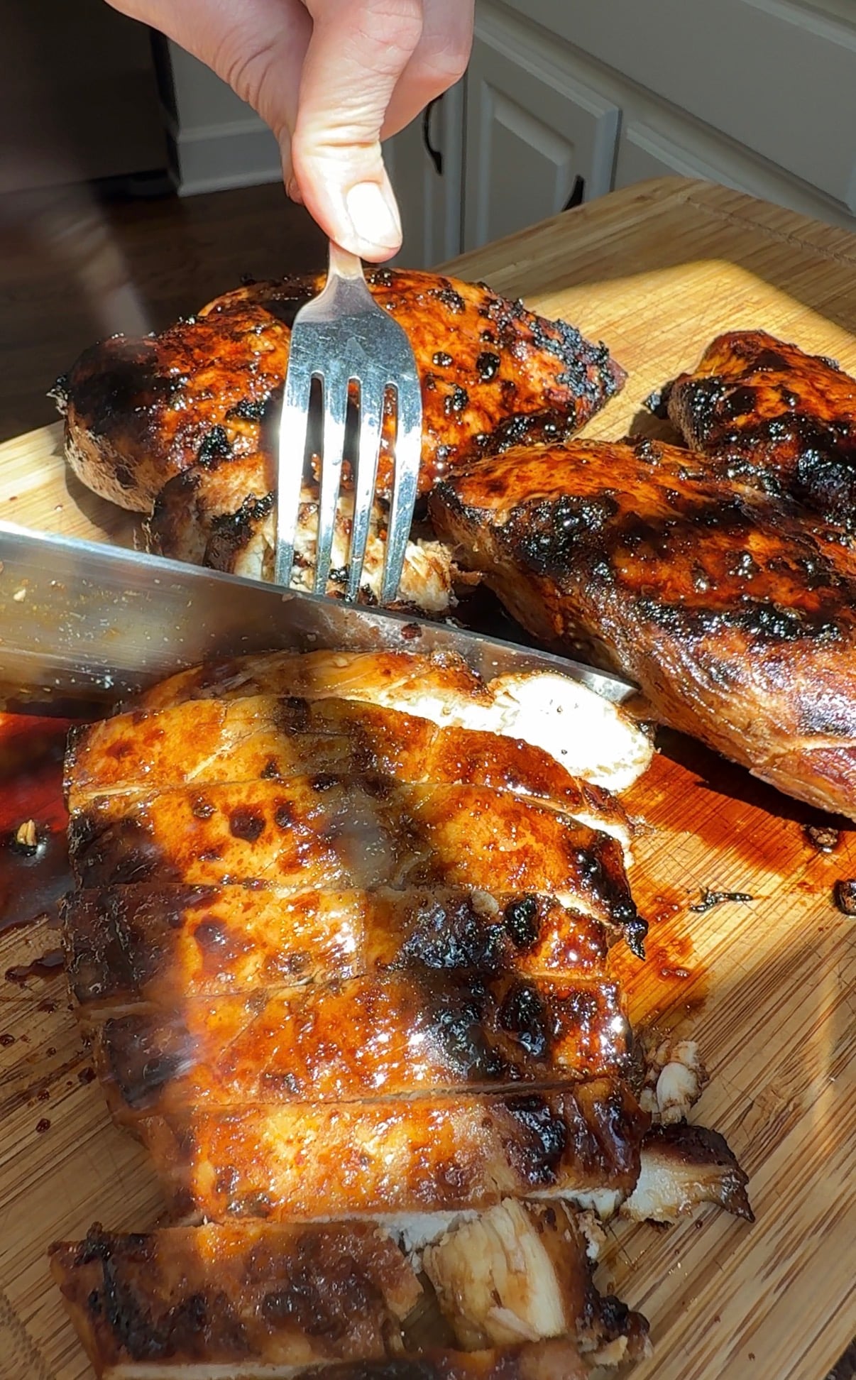 A hand uses a fork and knife to cut into glazed, grilled chicken breasts on a wooden cutting board in a bright kitchen.
