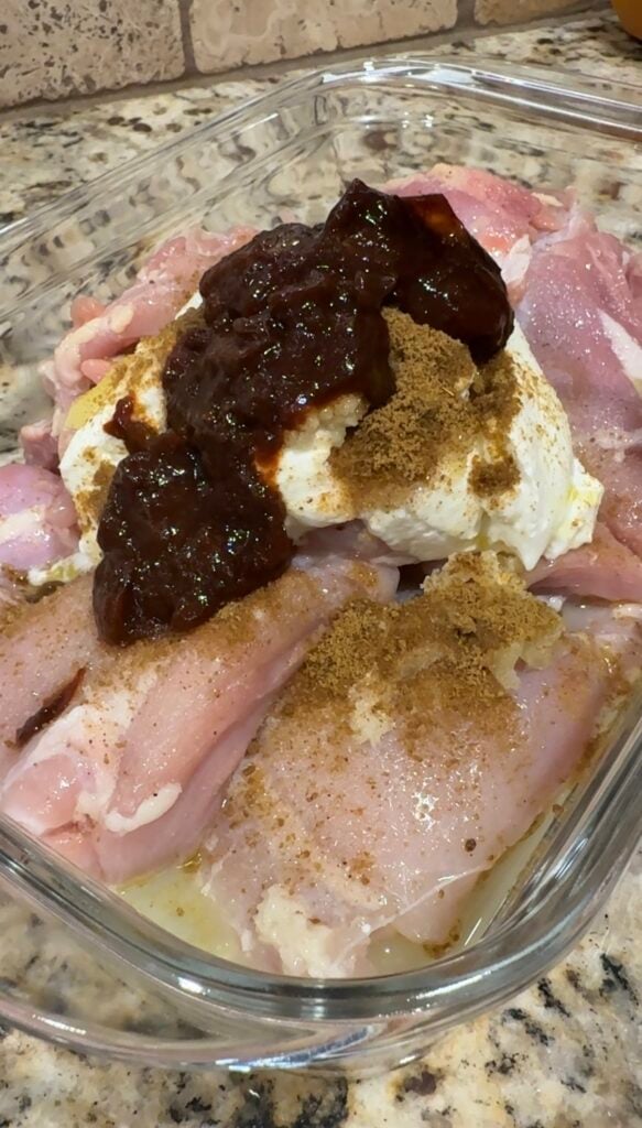 Raw chicken breasts in a glass dish topped with a dollop of white sauce, brown spice, and a dark red sauce, sitting on a granite countertop.
