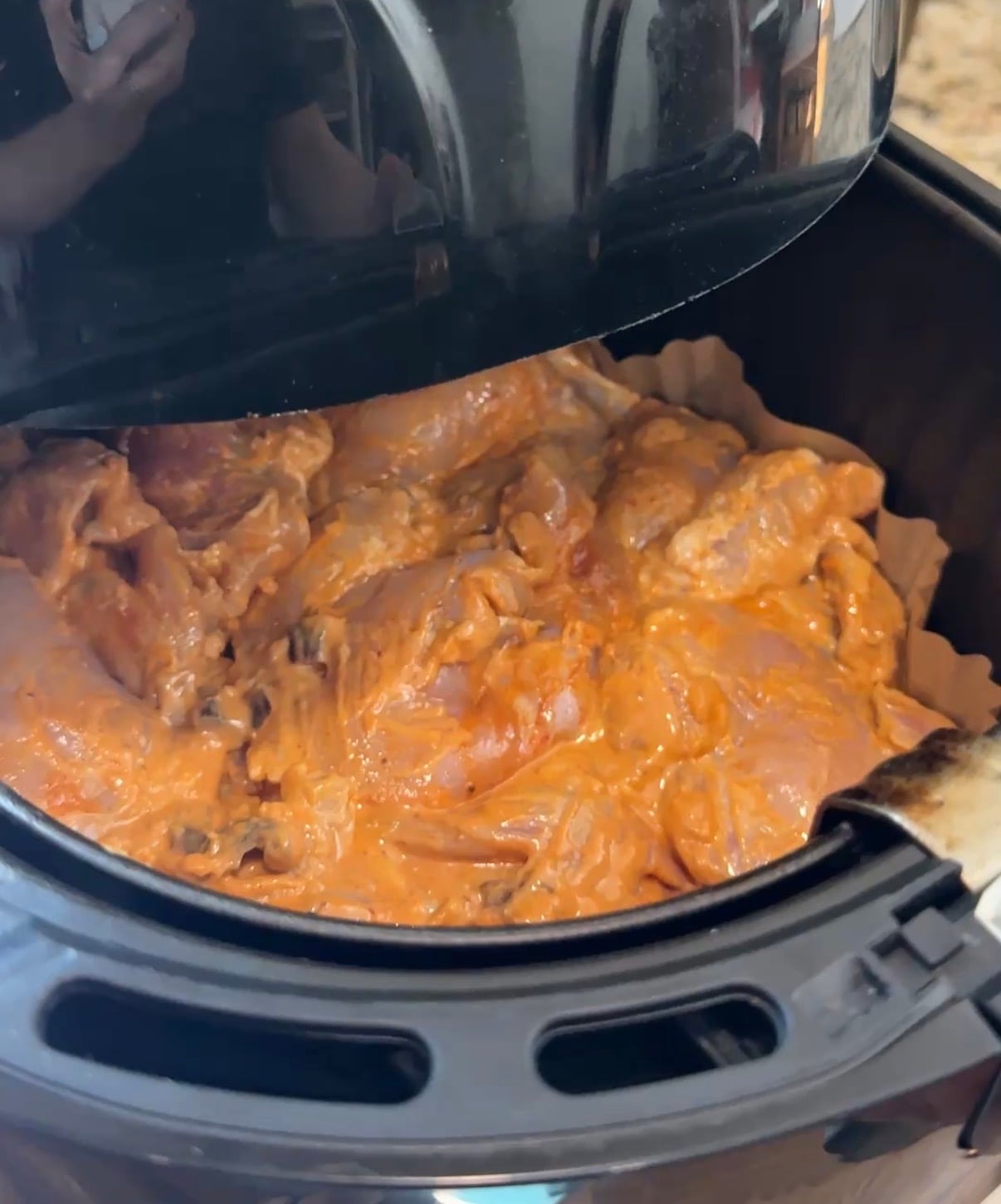 Marinated raw chicken pieces arranged on parchment paper inside an air fryer basket, with the air fryer lid partially open, ready for cooking.