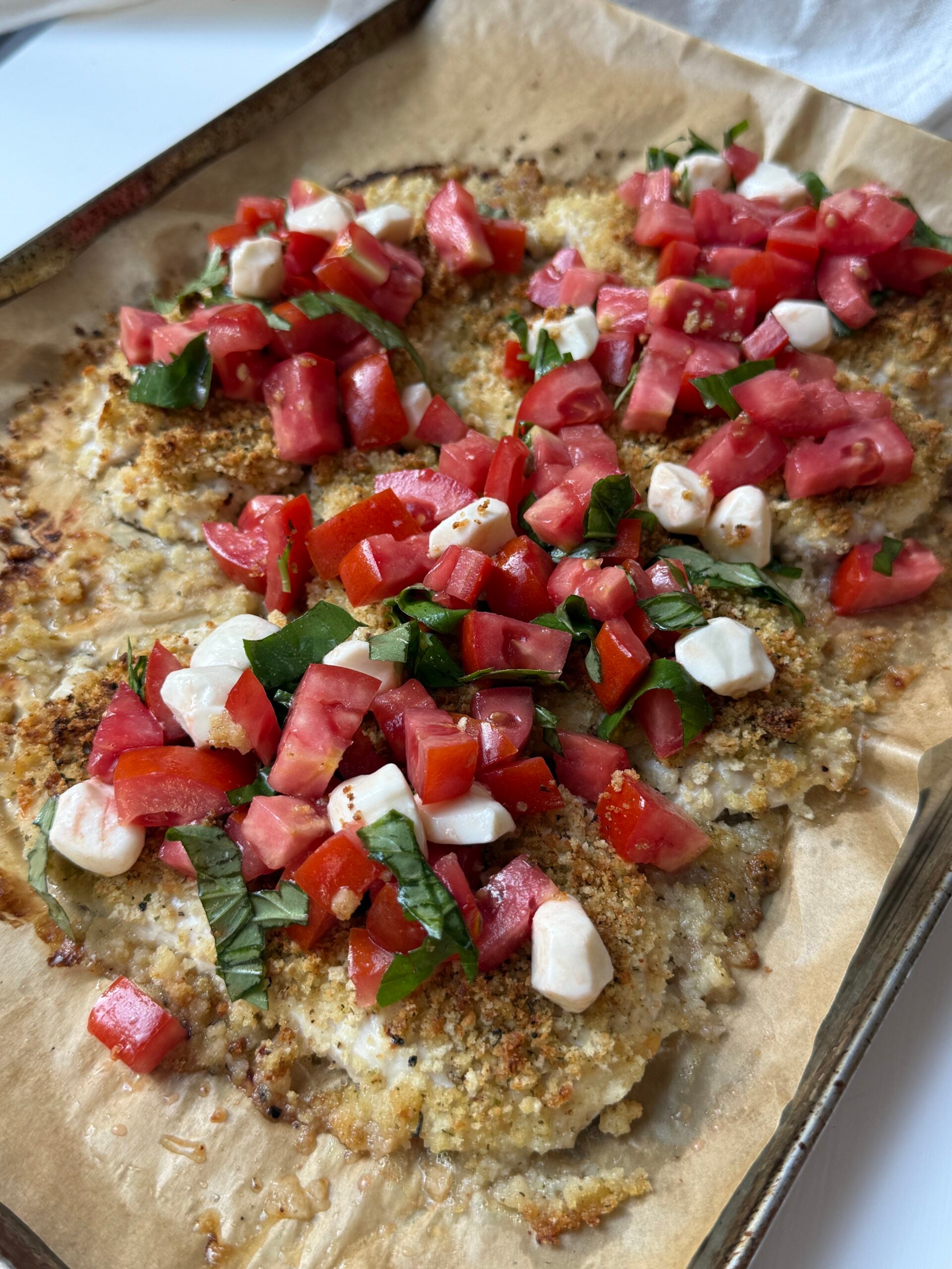 Breaded chicken on parchment paper, topped with diced tomatoes, mozzarella, and fresh basil, resembling a bruschetta-style presentation. The dish is colorful and served on a baking sheet.