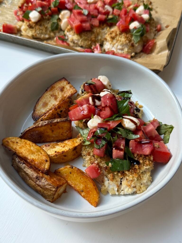 A bowl with breaded chicken topped with diced tomatoes, mozzarella, and basil, served with roasted potato wedges; similar food is on a tray in the background.