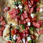 Breaded chicken topped with chopped tomatoes, mozzarella, fresh basil, and a drizzle of balsamic glaze on parchment paper.