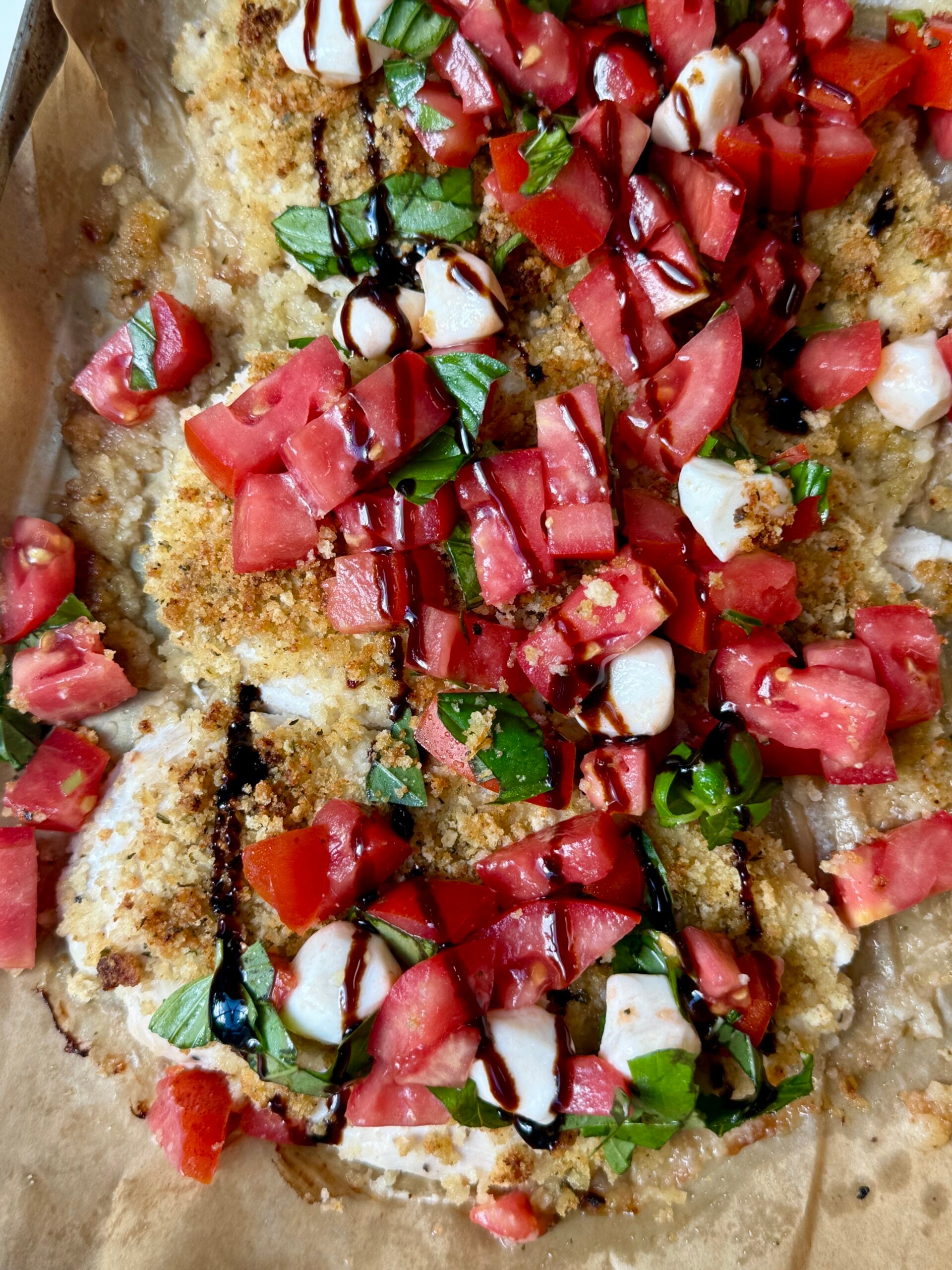 Breaded chicken topped with chopped tomatoes, mozzarella, fresh basil, and a drizzle of balsamic glaze on parchment paper.