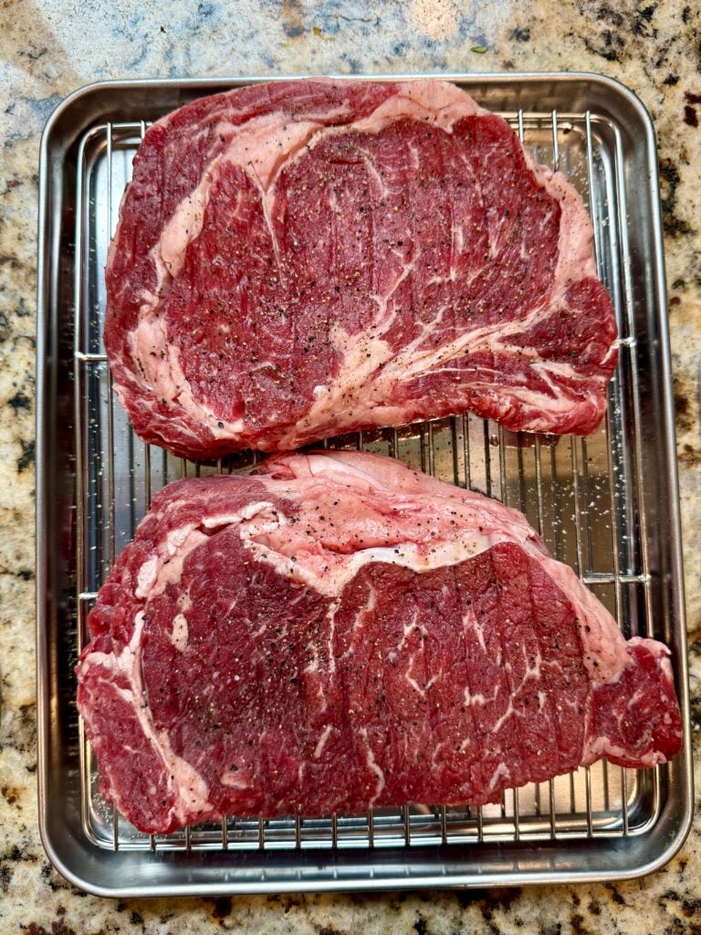 Two raw, marbled ribeye steaks seasoned with pepper sit on a wire rack in a metal tray, placed on a granite countertop.