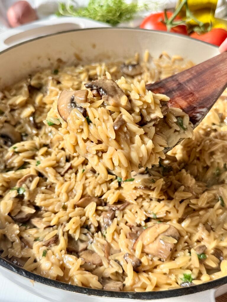 Creamy orzo pasta with sliced mushrooms and fresh herbs is cooked in a large white pan. Tomatoes, greens, and garlic are visible in the blurred background.