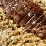 Sliced steak on a bed of creamy orzo pasta, garnished with grated cheese and herbs, served in a skillet.