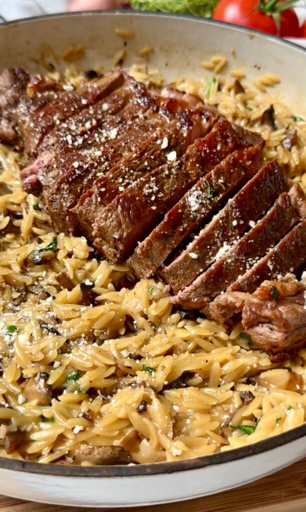Sliced steak on a bed of creamy orzo pasta, garnished with grated cheese and herbs, served in a skillet.