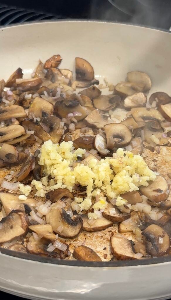 Sliced mushrooms and chopped shallots are being sautéed in a pan, with a pile of freshly minced garlic added on top, ready to be mixed in. Steam rises from the hot pan.