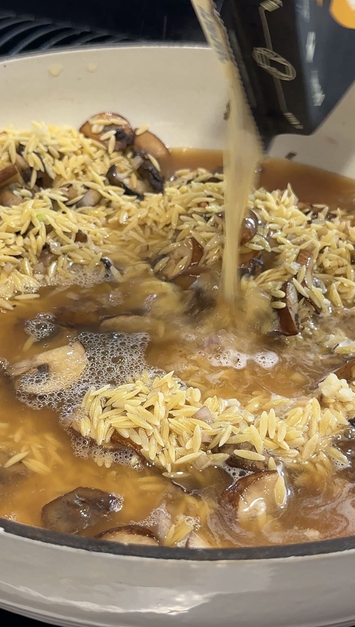 Orzo pasta and sliced mushrooms in a pan as broth is being poured in, creating a rich mixture with visible bubbles and grains starting to absorb the liquid.