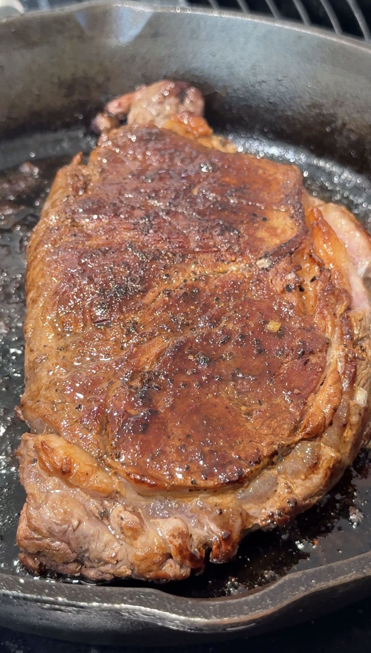 A well-seasoned steak sizzling in a cast iron skillet, with a browned, crispy crust and visible marbling, cooking juices, and oil bubbling around the edges.