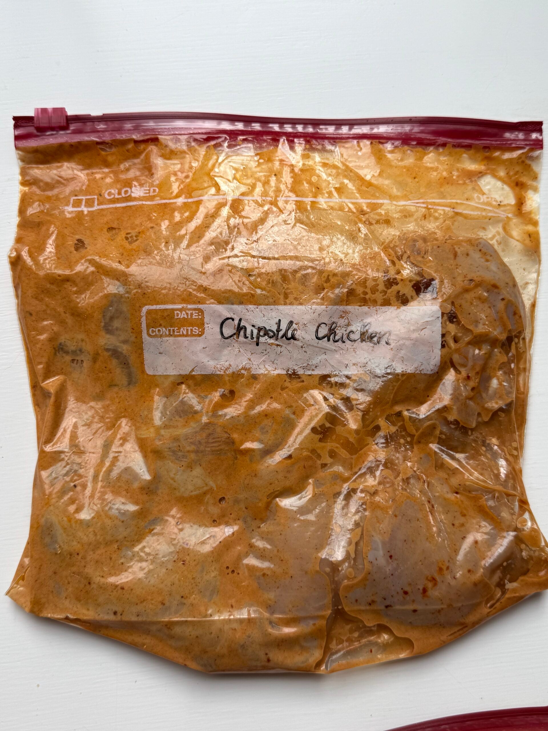 A clear zip-top bag filled with marinated chicken in an orange sauce. A white label on the bag reads “Chipotle Chicken” in handwritten text.