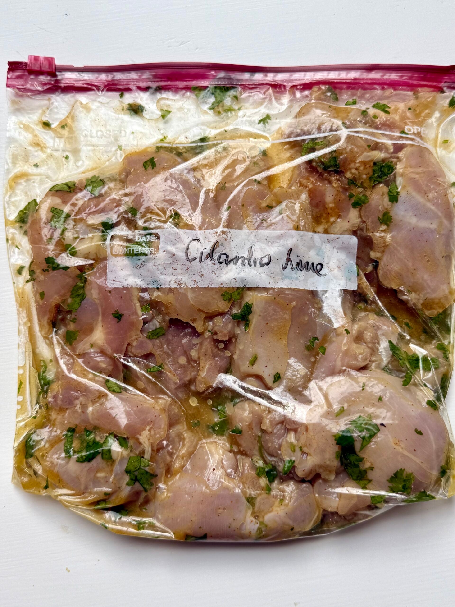 Raw chicken marinating in a clear ziplock bag labeled Cilantro Lime, with visible herbs and marinade surrounding the chicken pieces. The bag is placed on a white surface.
