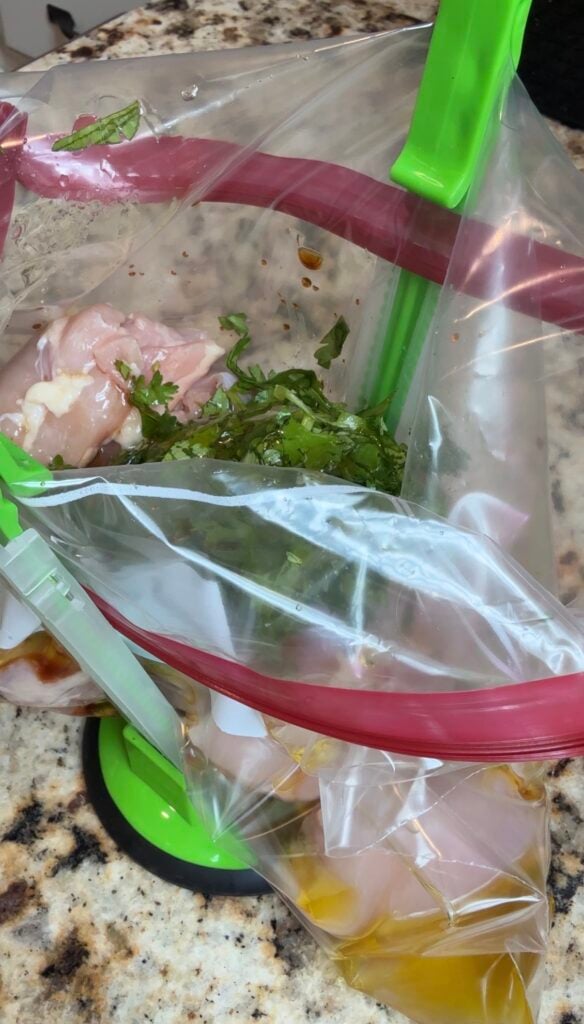 Raw chicken pieces, chopped cilantro, and marinade are in a zip-top plastic bag set open with a green bag holder on a granite countertop.