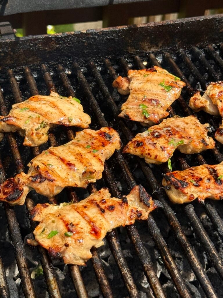 Grilled marinated chicken pieces cooking on a barbecue grill, with visible grill marks and some herbs sprinkled on the meat.