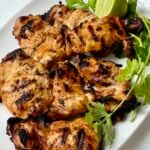 Grilled chicken pieces with char marks are served on a white plate, garnished with fresh cilantro and a halved lime.
