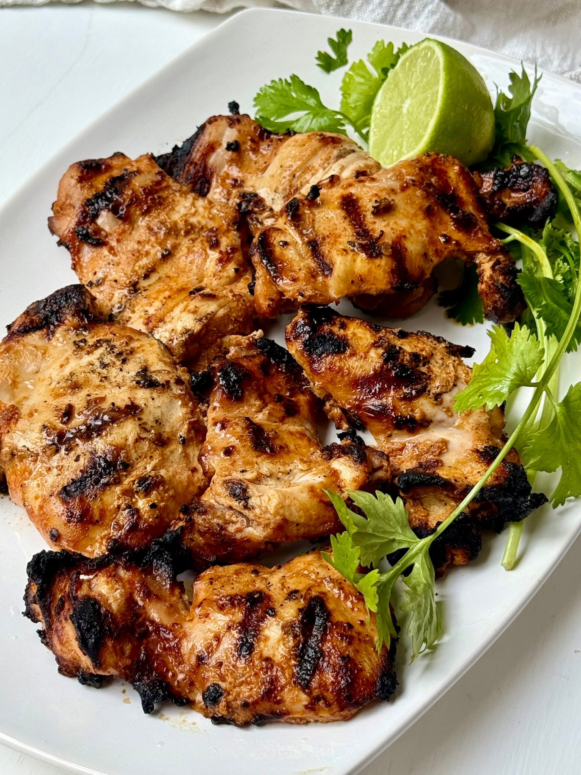 Grilled chicken pieces with char marks are served on a white plate, garnished with fresh cilantro and a halved lime.