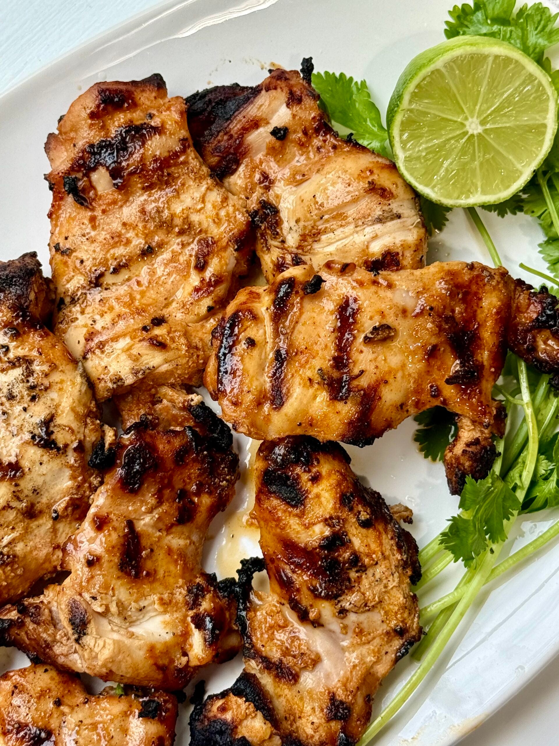 Grilled chicken pieces with visible char marks on a white plate, garnished with fresh cilantro and a sliced lime.