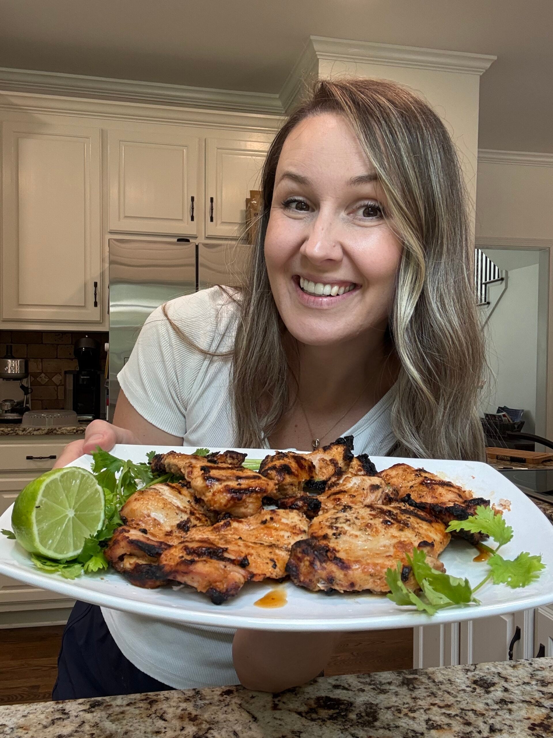 A smiling woman in a kitchen holds a plate of grilled chicken garnished with cilantro and lime.