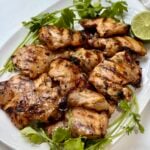 Grilled chicken pieces on a white plate, garnished with fresh cilantro and a halved lime.