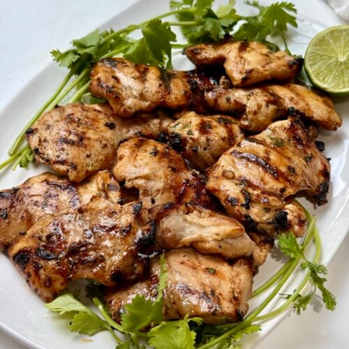 Grilled chicken pieces on a white plate, garnished with fresh cilantro and a halved lime.