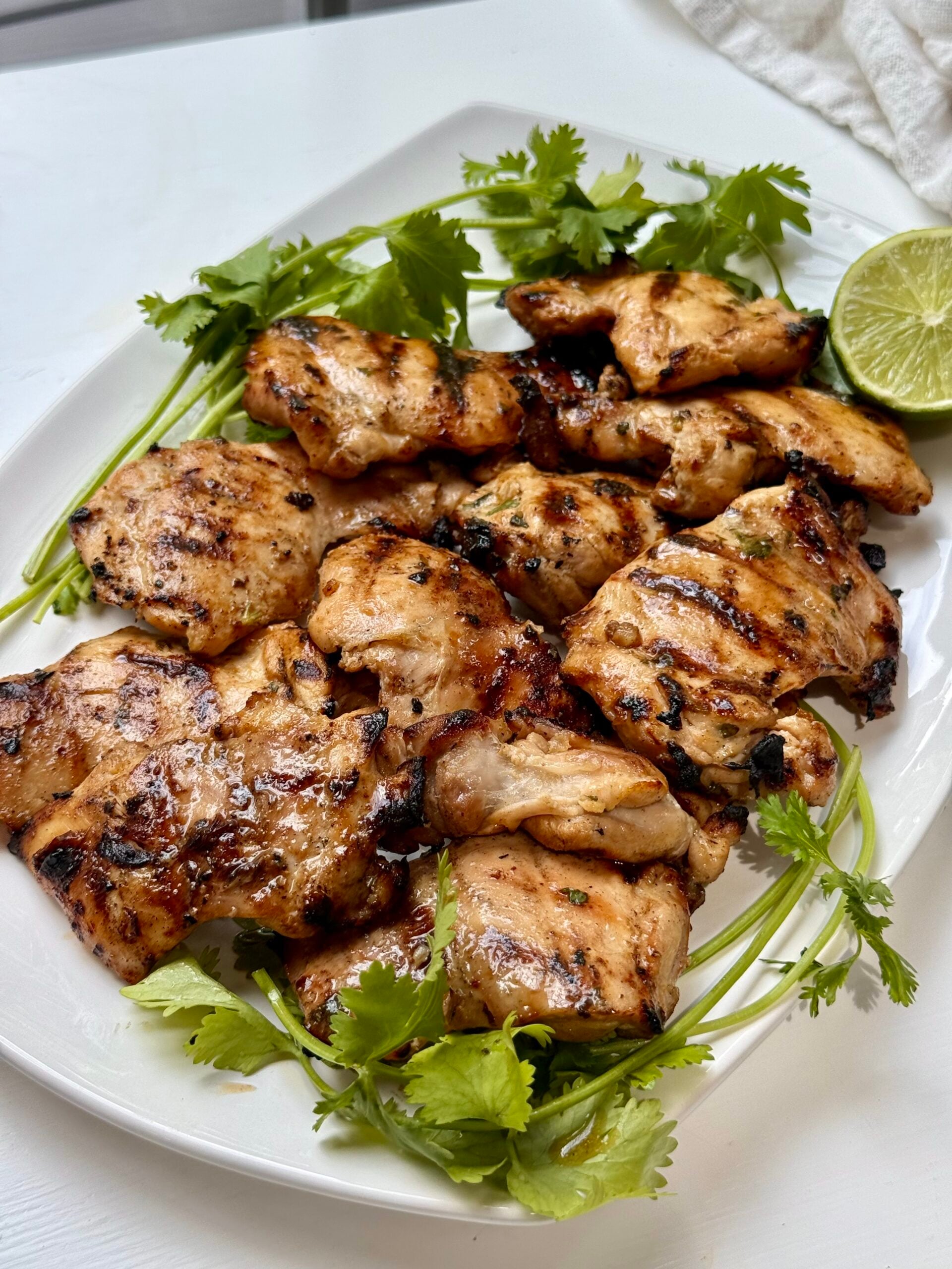 Grilled chicken pieces on a white plate, garnished with fresh cilantro and a halved lime.