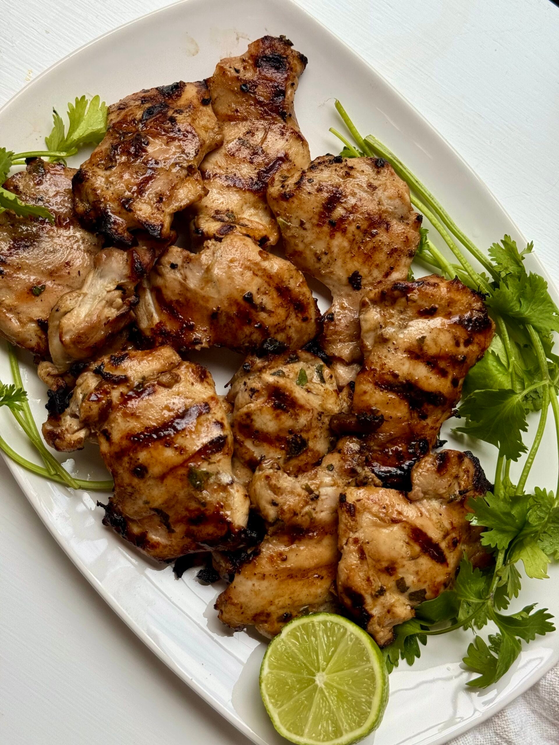 Grilled chicken thighs arranged on a white plate with grill marks, garnished with fresh cilantro and a halved lime.