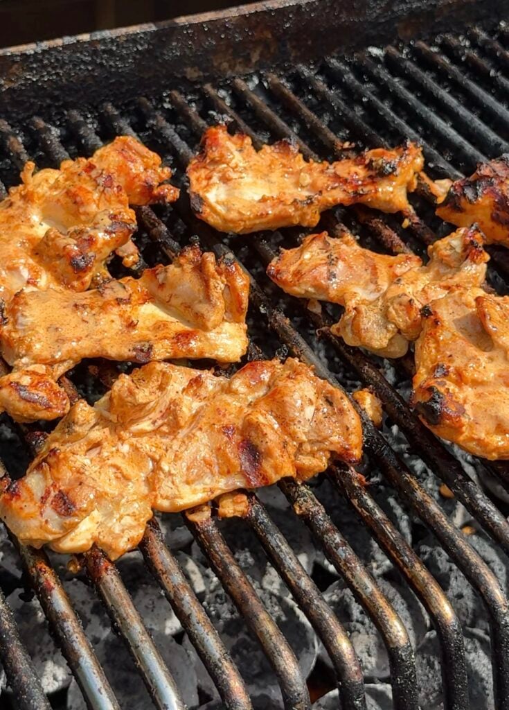 Marinated pieces of chicken are grilling on a barbecue, with char marks visible on the meat and grill grates over hot coals.