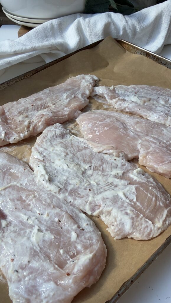 Raw chicken cutlets coated with a white marinade, spread out on a parchment-lined baking sheet, ready to be cooked.