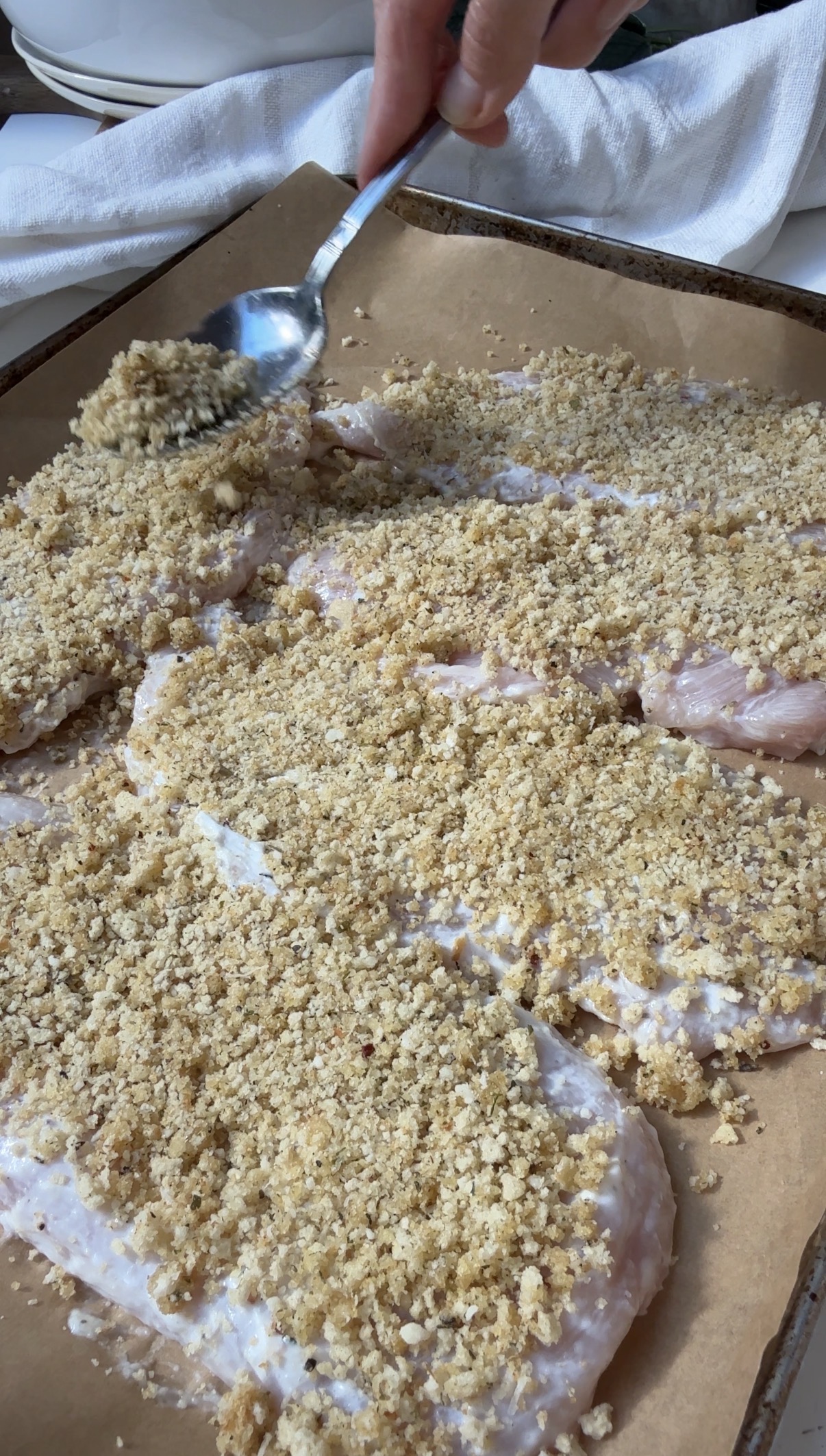 A hand sprinkles breadcrumb topping from a spoon onto several raw, breaded chicken breasts arranged on a baking sheet lined with parchment paper.