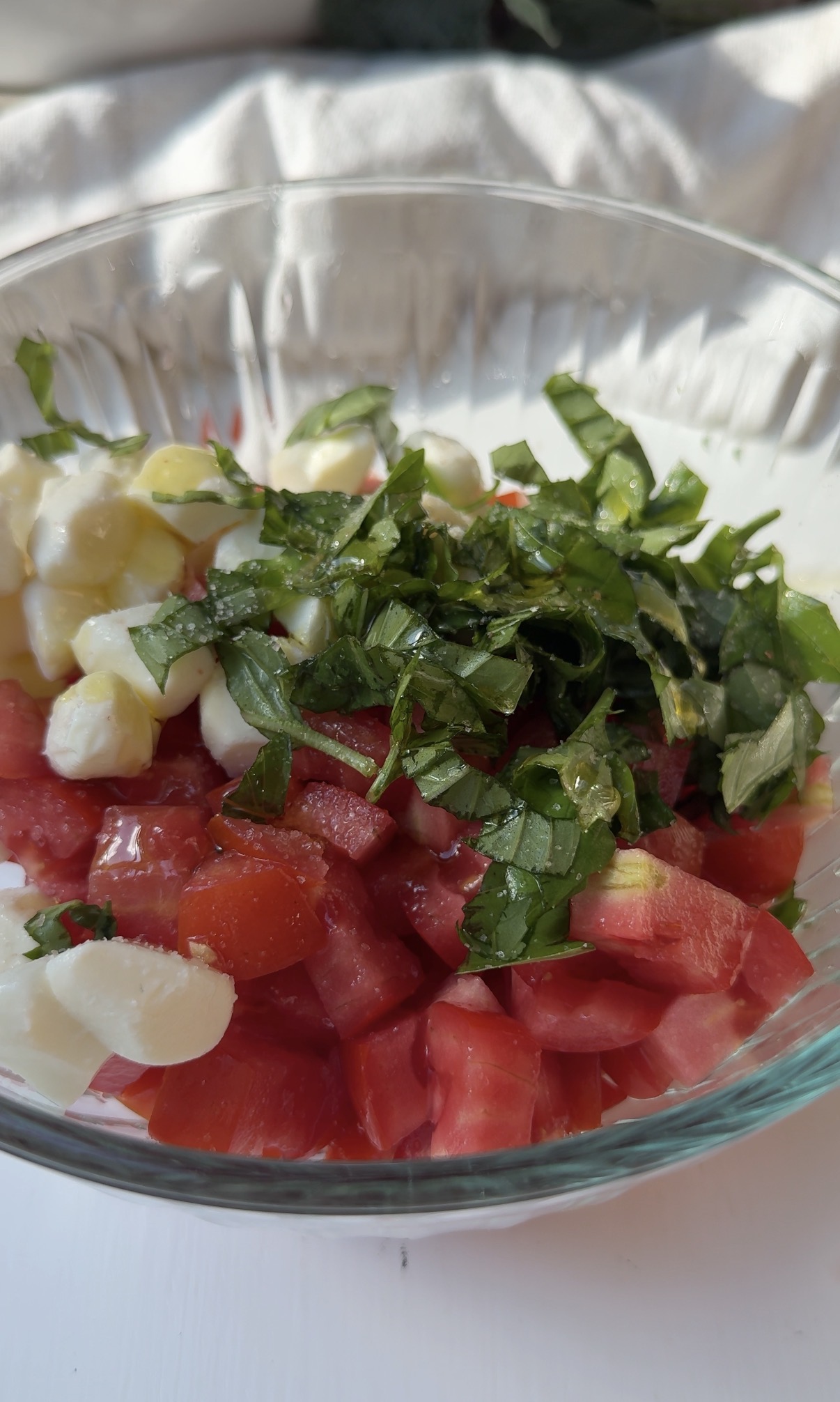A glass bowl filled with chopped tomatoes, mozzarella cheese, and fresh basil leaves, placed on a white surface with soft light.