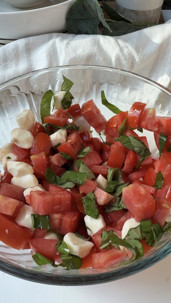 A glass bowl filled with chopped tomatoes, mozzarella cheese, and fresh basil leaves, placed on a white surface with soft light.