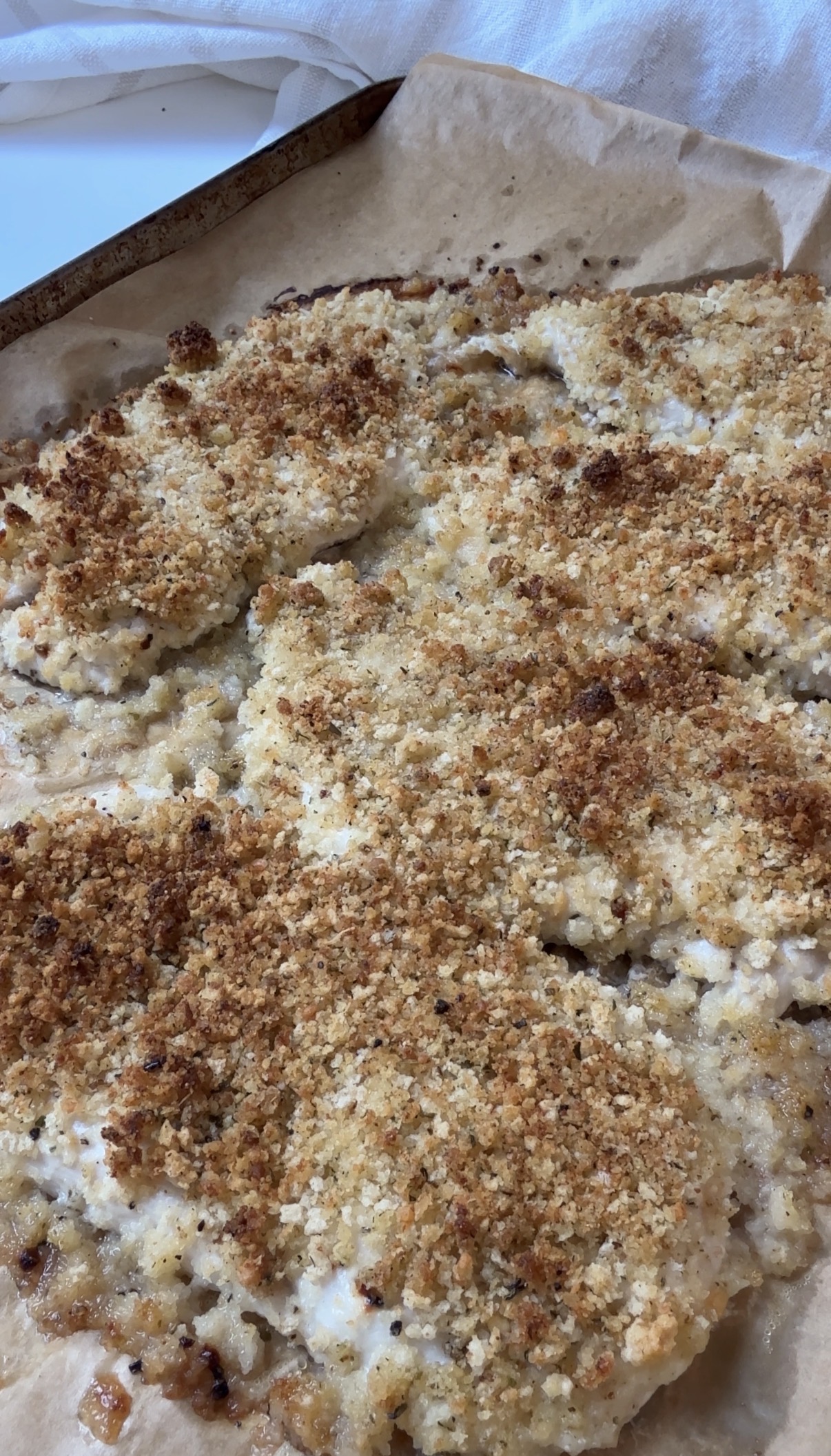 A close-up of a baked dish with a golden-brown breadcrumb topping on parchment paper, likely breaded chicken or fish, showing a crispy texture.