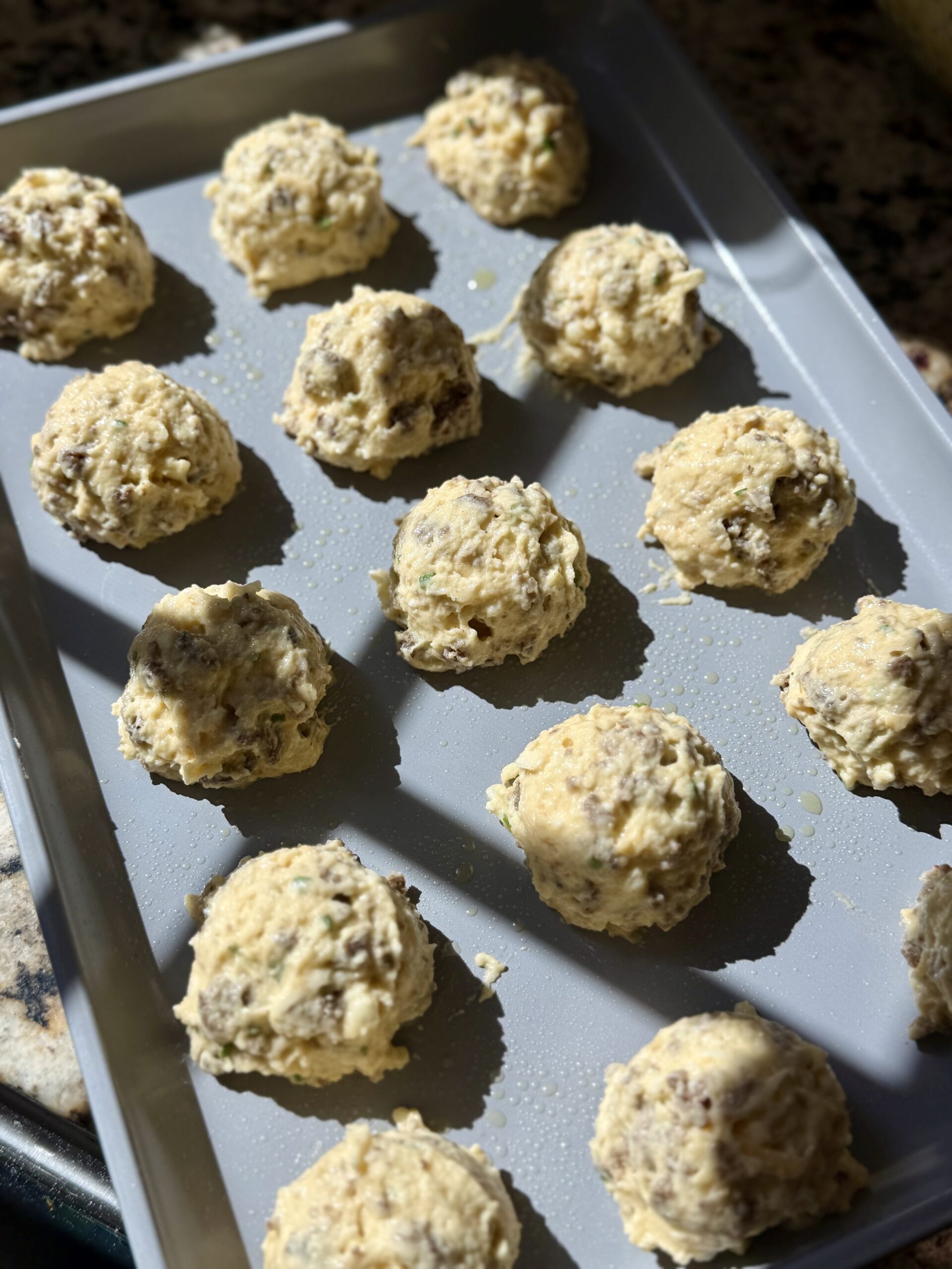 A baking tray holds rows of unbaked sausage and cheese balls, spaced evenly and ready to go into the oven. Sunlight shines on the tray from one side.