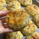 A hand holds a cheesy, golden-brown biscuit with herbs and bits of sausage, with more similar biscuits in a baking tray in the background.